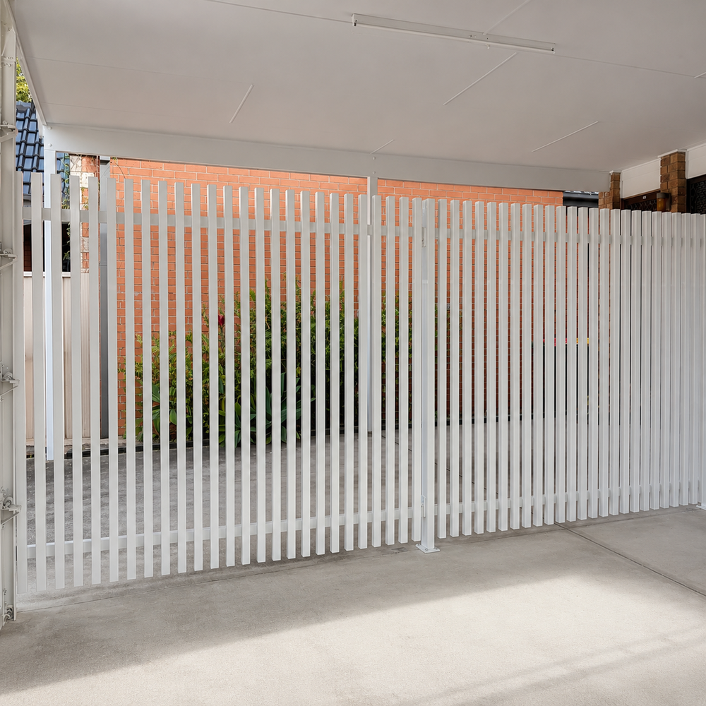 A white vertical batten fence installed along the side of a covered patio or carport, with a brick wall and greenery visible in the background