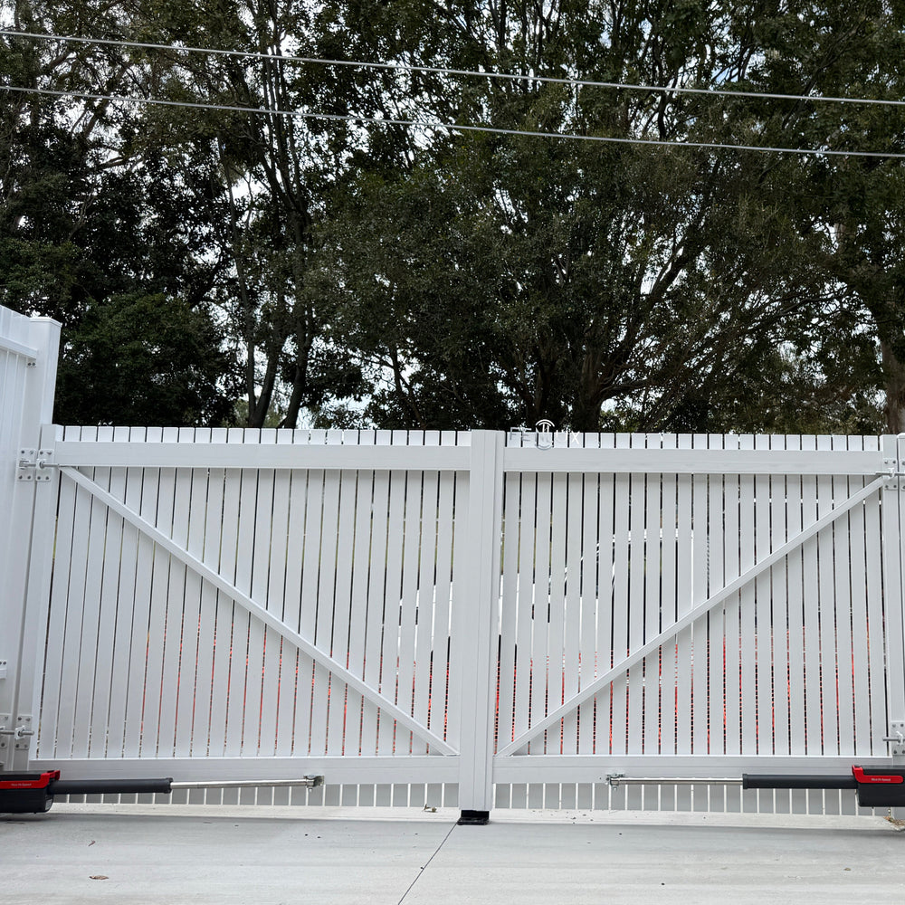 White vertical picket swing gate with white fence, surrounded by trees and a clear sky.