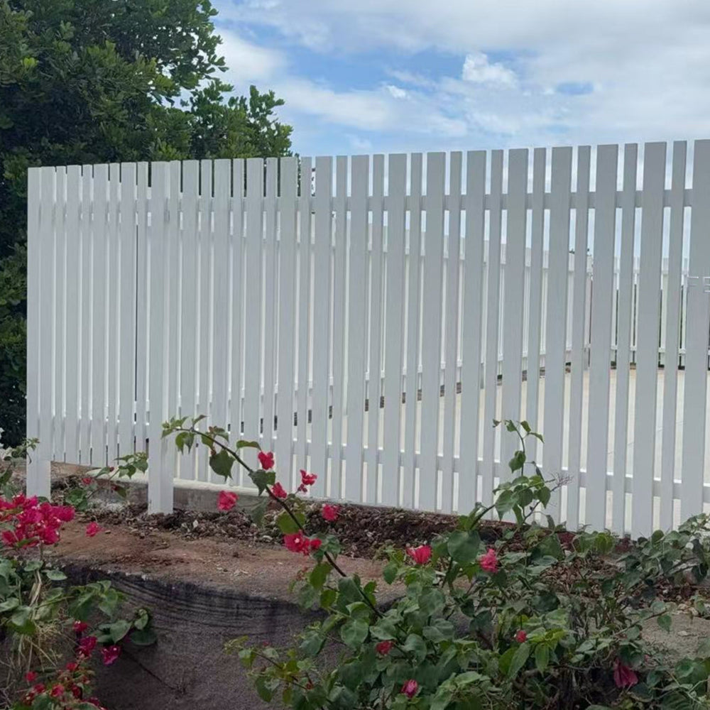 White combo picket fence with flowers and a bench in front, blue sky with clouds in the background