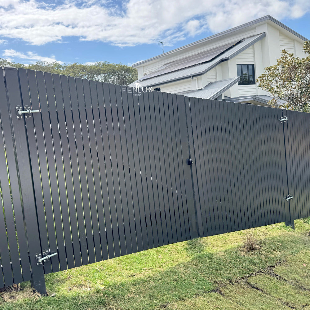 Black aluminium vertical picket swing gate in front of a house with a blue sky and clouds.