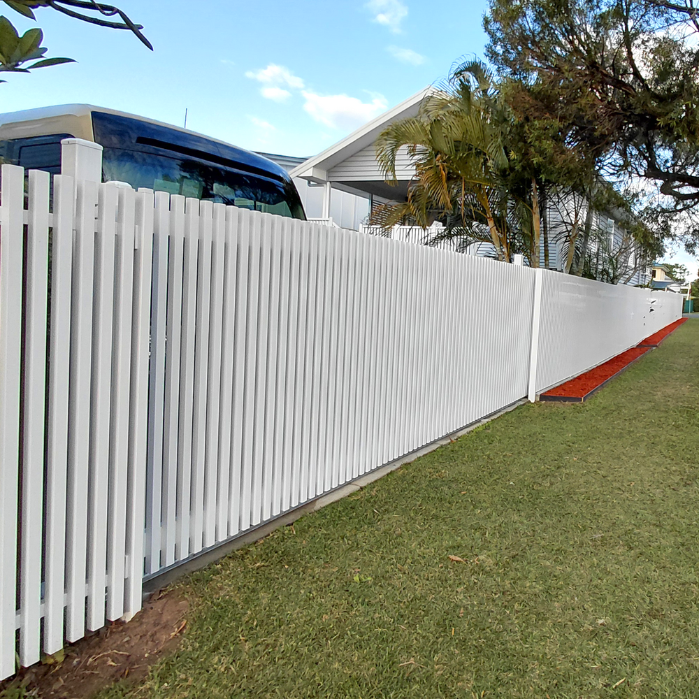 A long white batten fence installed along the side of a house, with green grass in front and trees and a blue sky in the background.